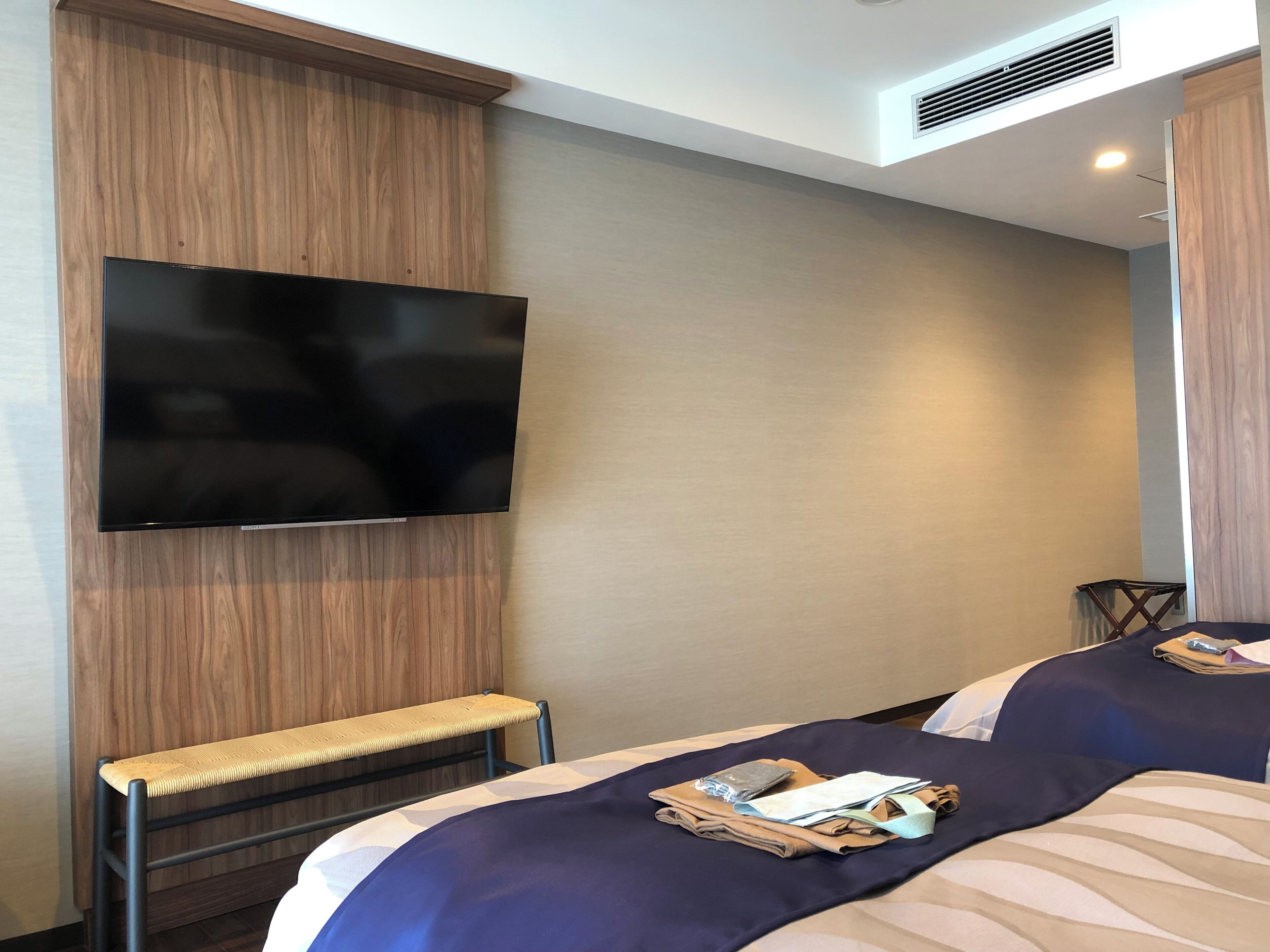 Una camera d'albergo con una televisione a schermo piatto montata a parete, una panca e due letti con asciugamani ordinatamente piegati e una borsa.