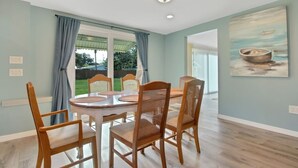 Un área de comedor con una mesa redonda y seis sillas de madera, ventanas amplias con cortinas azules, y un cuadro de un barco en la pared.