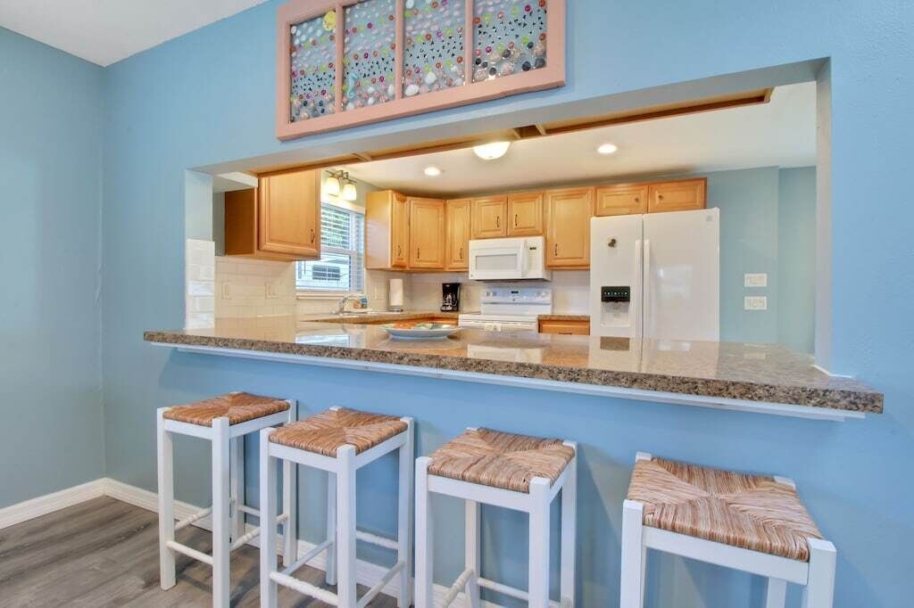 Una cocina con gabinetes de madera, electrodomésticos blancos y encimera de granito. Hay tres taburetes blancos con asientos de mimbre.