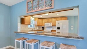 Una cocina con gabinetes de madera, electrodomésticos blancos y encimera de granito. Hay tres taburetes blancos con asientos de mimbre.