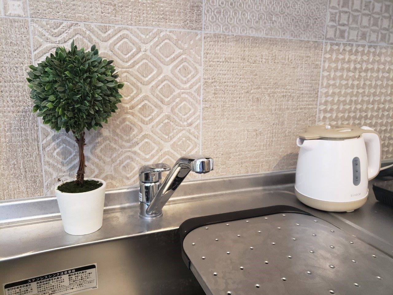 A kitchen with a sink, a potted plant, a kettle, and a patterned backsplash.