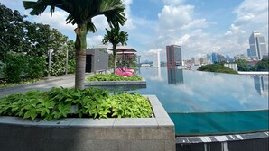 An infinity pool with a city skyline in the background, surrounded by greenery and palm trees. - Opus Residences (Kuala Lumpur)