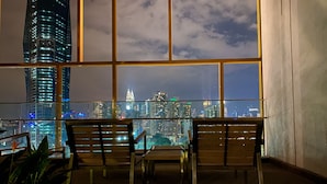 Ein Zimmer mit großem Fenster und Blick auf eine beleuchtete Stadtlandschaft bei Nacht. Im Vordergrund befinden sich zwei Holzstühle und ein kleiner Tisch.
