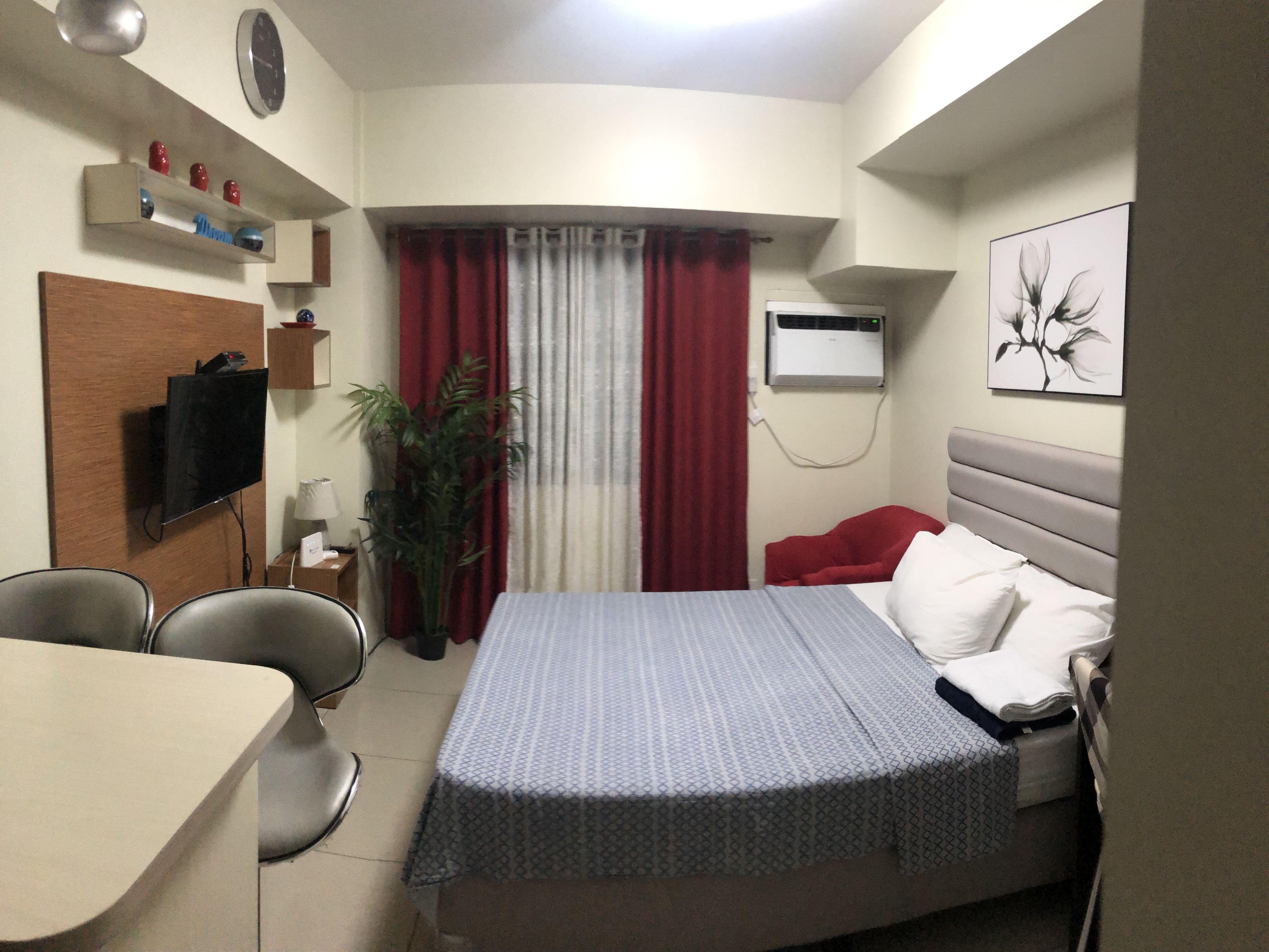 Habitación de hotel con cama, escritorio, silla, televisor, planta y un cuadro en la pared.