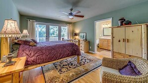 A bedroom with a bed, a nightstand, a lamp, a ceiling fan, a wicker chair, and a view of the kitchen. - Exclusive private mountain and lake vacation in the Cohutta wilderness (Ellijay)