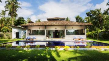 Une maison moderne à deux étages, dotée d’une piscine à l’avant, et entourée d’une végétation luxuriante et de palmiers.