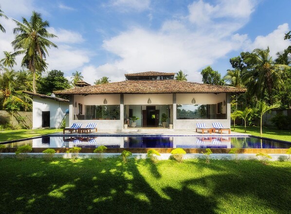 Una casa moderna de dos pisos con piscina frente a ella, rodeada de exuberante follaje y palmeras.