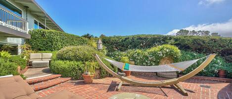 Un patio con una hamaca, mobiliario exterior y vistas a un jardín exuberante y colinas lejanas.