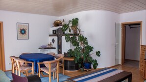 Una sala de estar acogedora con una mesa de comedor, sillas, un sofá y una mesita de centro. Hay una planta en maceta y una estantería decorativa con diversos objetos.