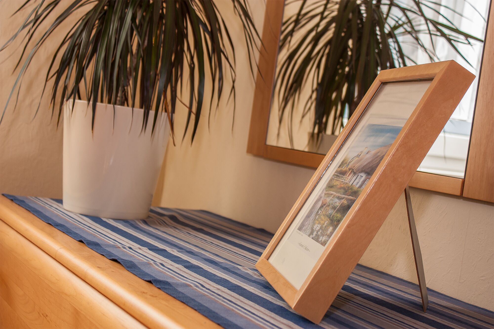 Un tableau encadré appuyé contre un mur, une plante en pot et un tissu à rayures posé sur une surface en bois.