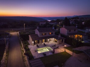 A stone house with a swimming pool, outdoor seating, and a view of the surrounding landscape at dusk.