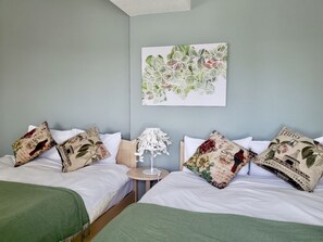 Habitación de hotel con dos camas, almohadas estampadas con motivos florales, una mesita con una lámpara y una obra de arte floral en la pared.
