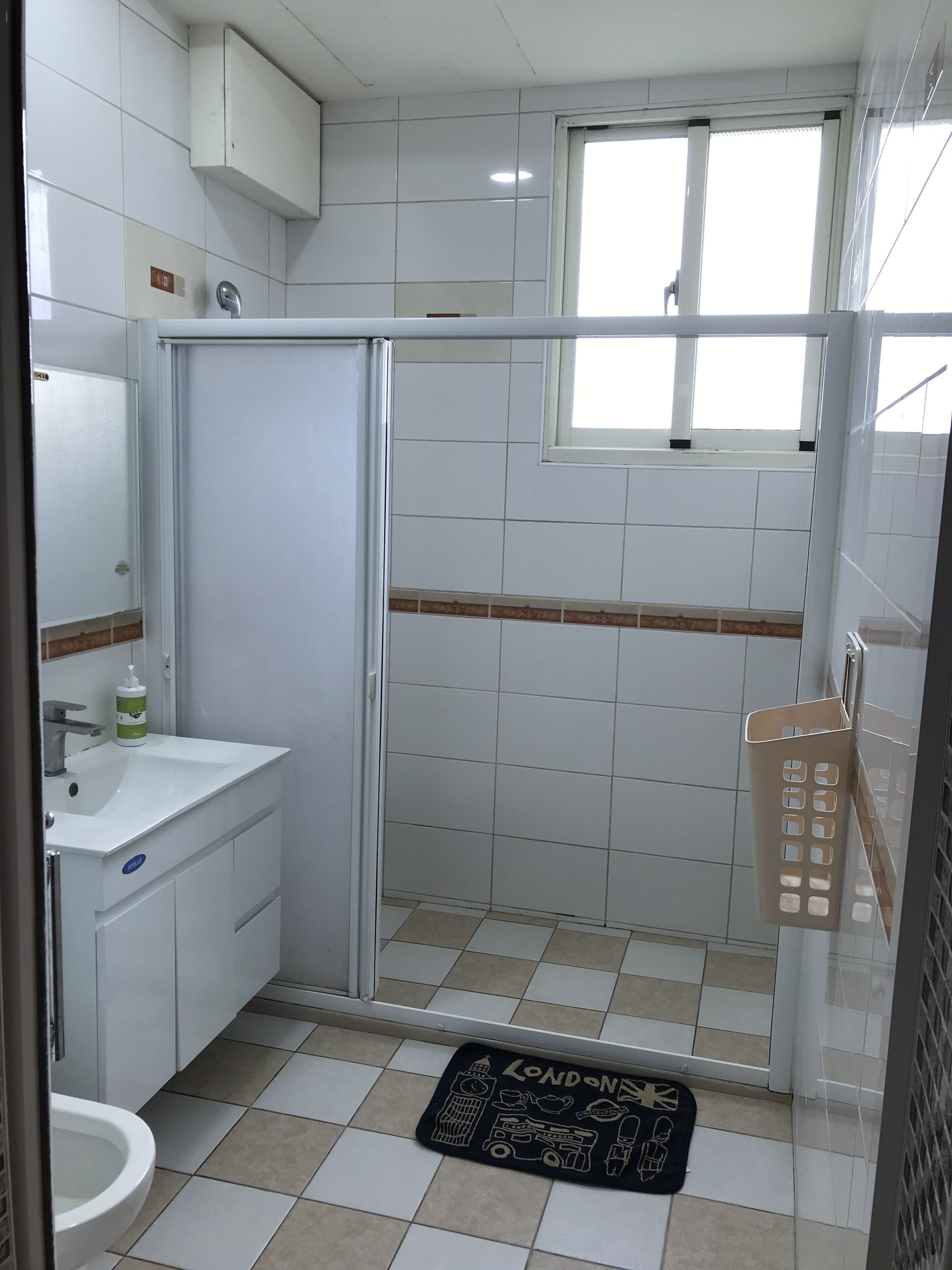 A bathroom with a sink, mirror, and shower. There is a window above the shower, and a mat with London landmarks on the floor.