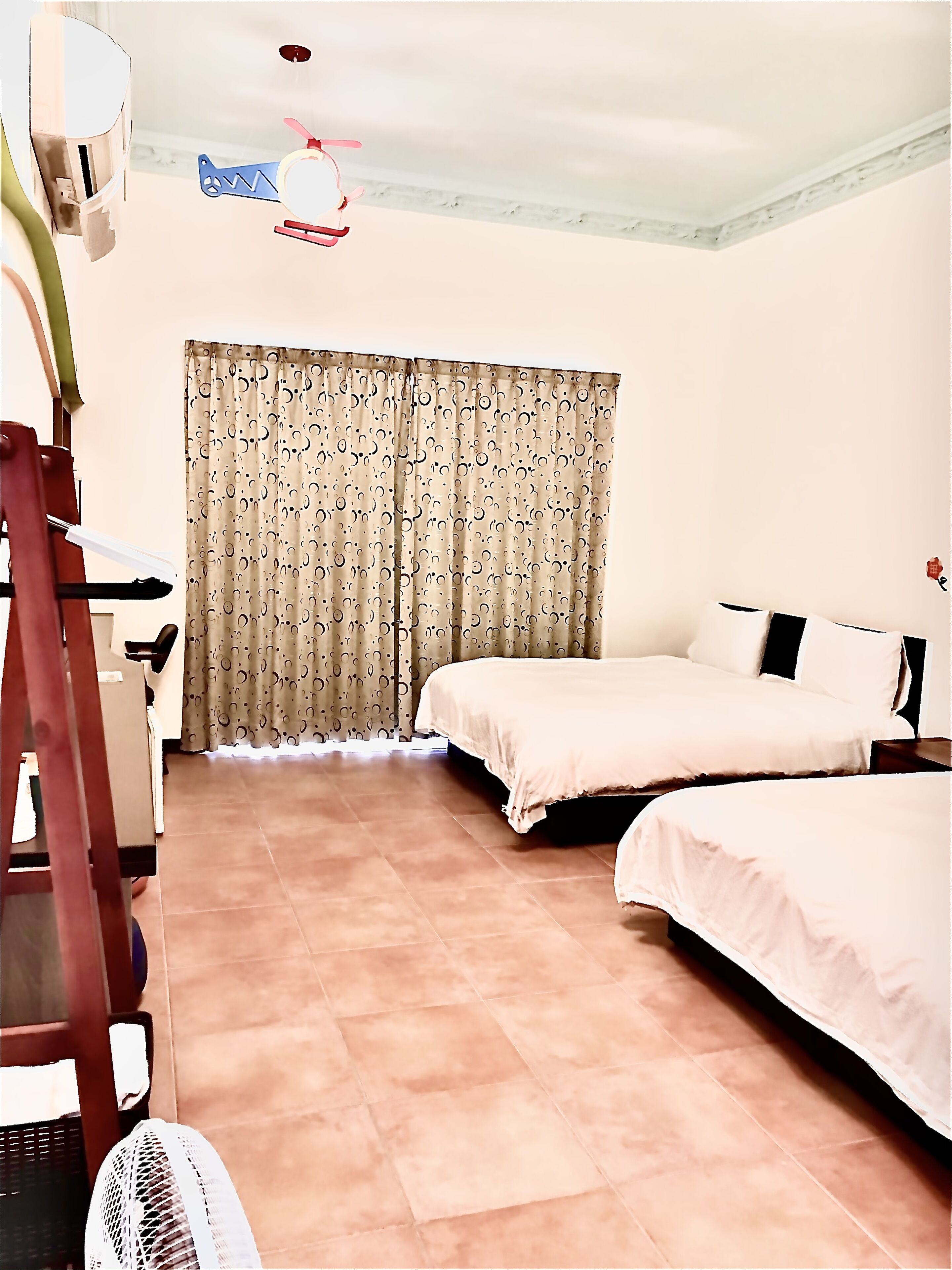 A hotel room with two beds, a desk, a chair, a fan, and a curtain with a circular pattern.