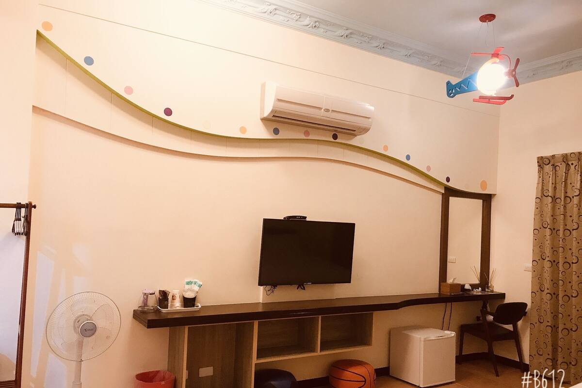 A room with a flat-screen TV mounted on the wall, a standing fan, a basketball, a small white refrigerator, and a desk with a chair.
