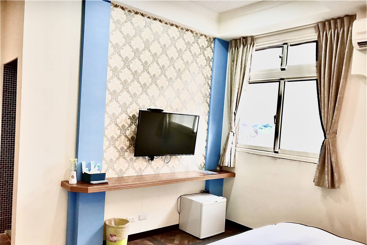 A hotel room with a bed, a TV mounted on the wall, a small air conditioner, and a window with curtains.