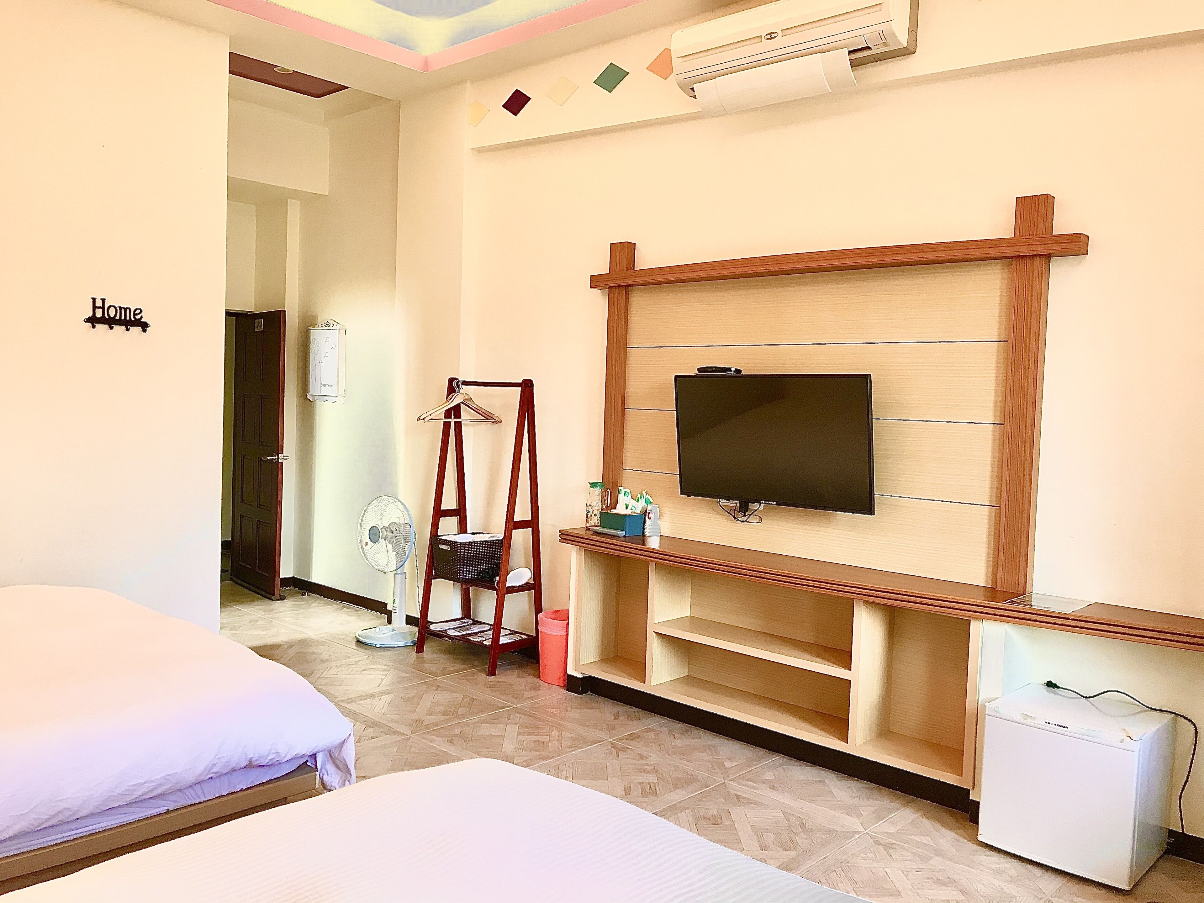 A hotel room with a bed, a TV mounted on the wall, a small fridge, and a wooden shelf.