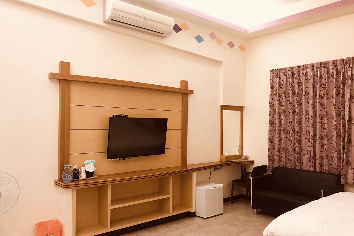 A hotel room with a flat-screen TV mounted on a wooden shelf, a small refrigerator, a desk with a mirror, a black chair, and a bed with white bedding.