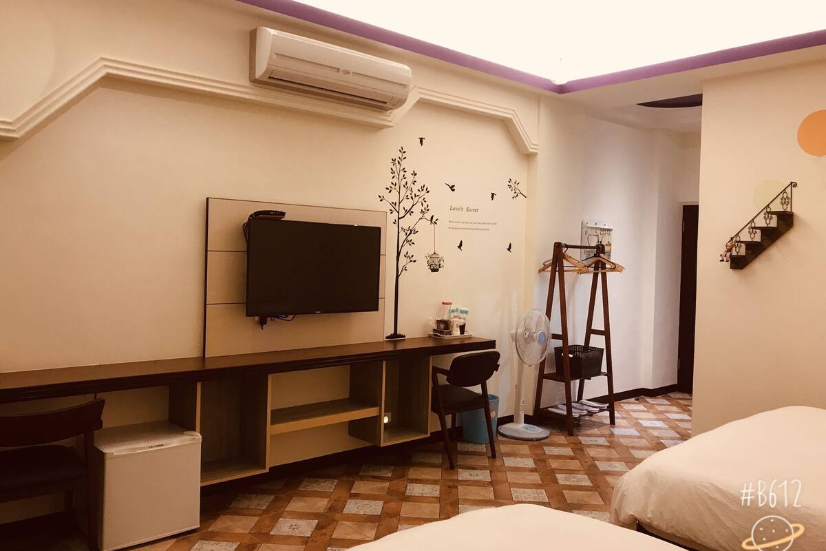 A hotel room with a flat-screen TV, a desk with a chair, a small refrigerator, a standing fan, and a bed.