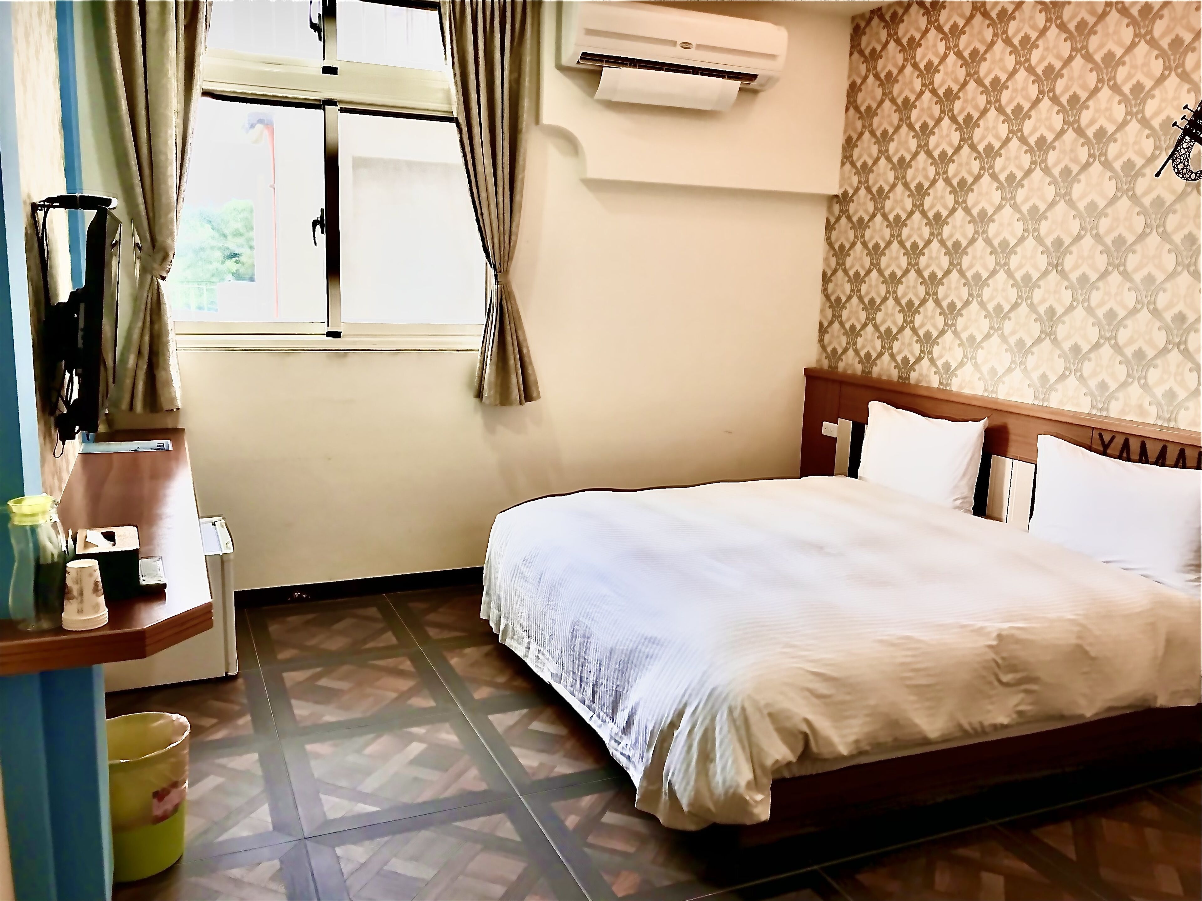 A hotel room with a bed, a desk, an air conditioning unit, and a patterned wall.