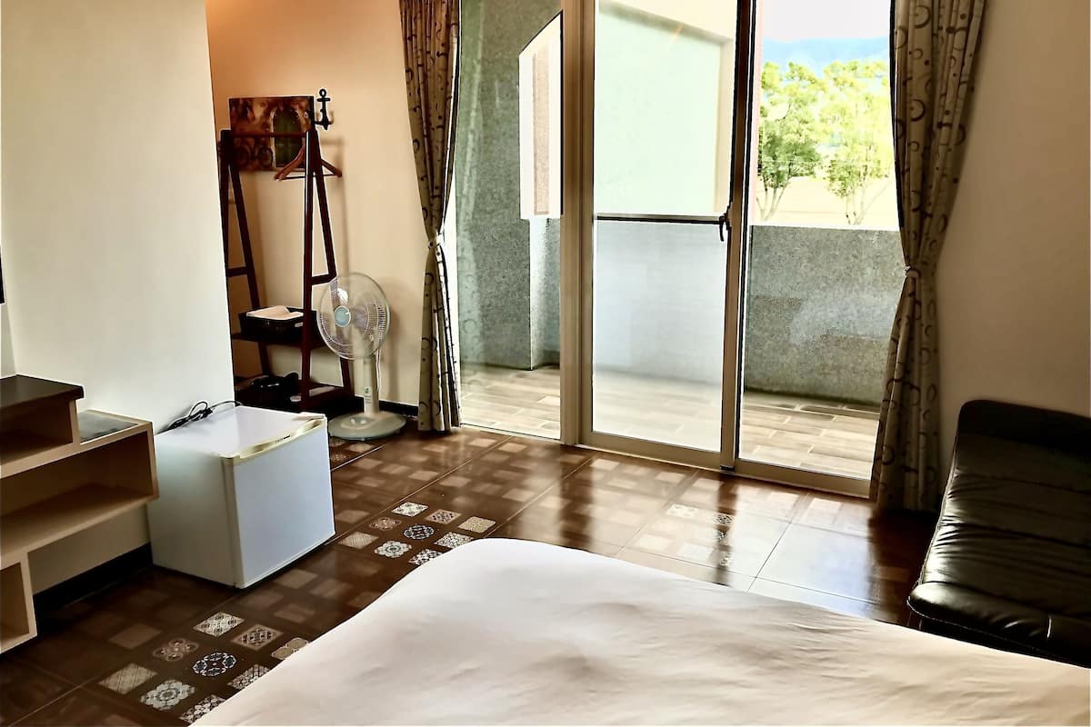 A hotel room with a bed, a desk, a chair, a fan, and a view of the outdoors through a sliding door.