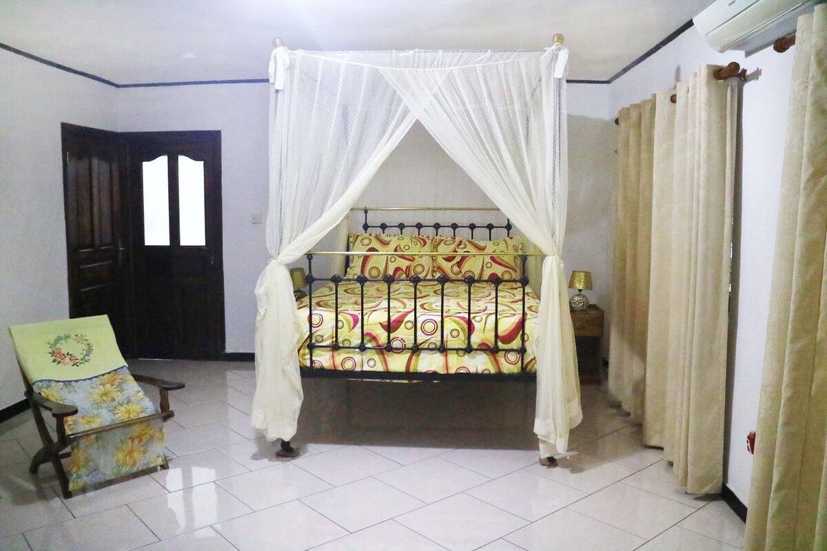 Una camera da letto con un letto a baldacchino, una poltrona con cuscino fiorito e un comodino con lampada.