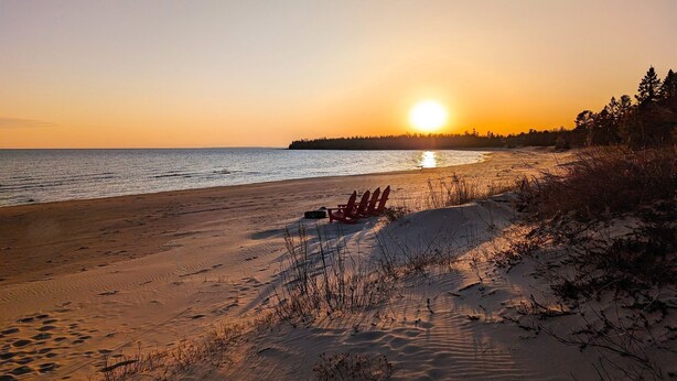 A beach with a row of red chairs facing the ocean, a clear sky, and the sun setting or rising. - Cedar Loft on Lake Michigan (Naubinway)
