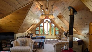 A cozy attic room with a sloped ceiling, a large window, a red stove, and a television. - Cedar Loft on Lake Michigan (Naubinway)