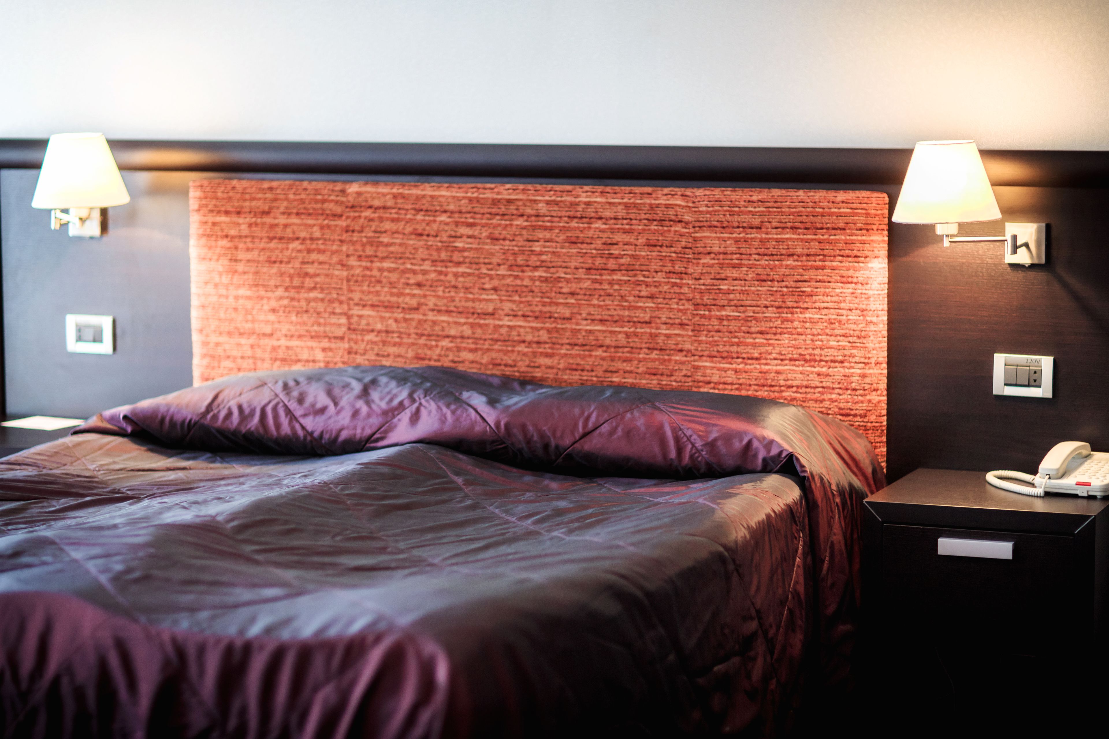 A hotel room with a bed, two bedside lamps, a nightstand with a phone, and a textured headboard.