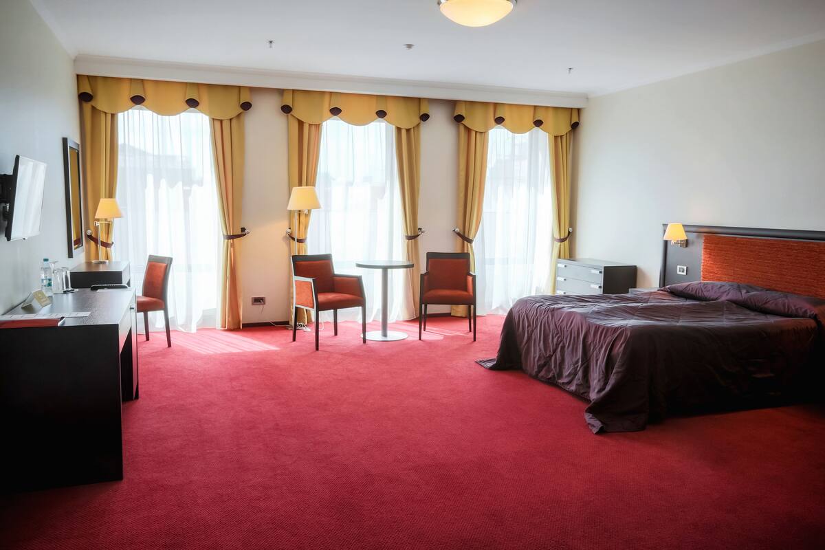 Habitación de hotel con alfombra roja, dos camas individuales, un escritorio con silla, una mesita con dos sillas y ventanas amplias con cortinas amarillas.