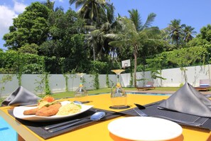 A dining table with a plate of food, cutlery, and a glass of drink, set against a backdrop of a pool and lush greenery. - Serenity Tissa (Thissamaharama)