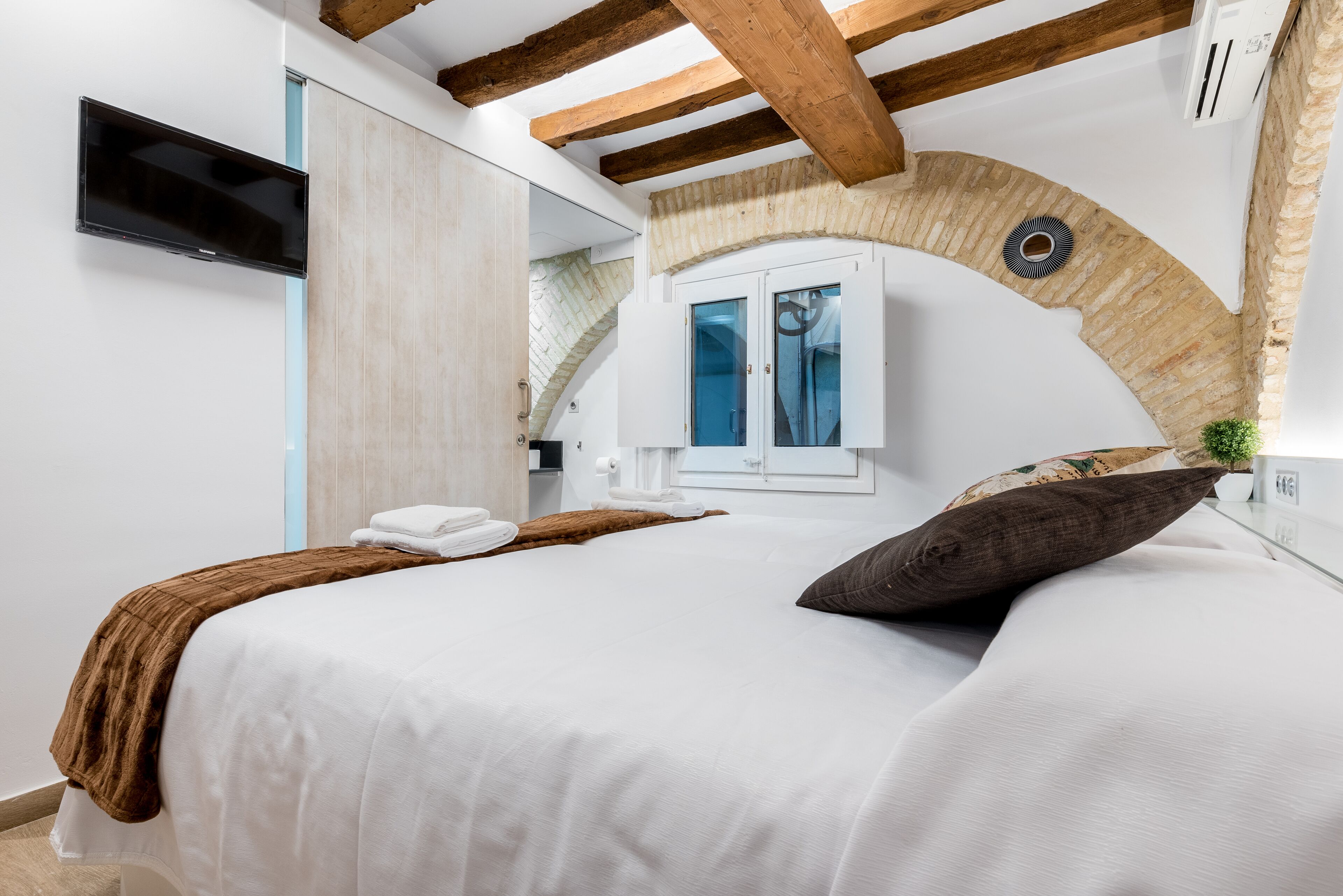 A bedroom with a bed, a TV, a window, and a wooden ceiling beam.