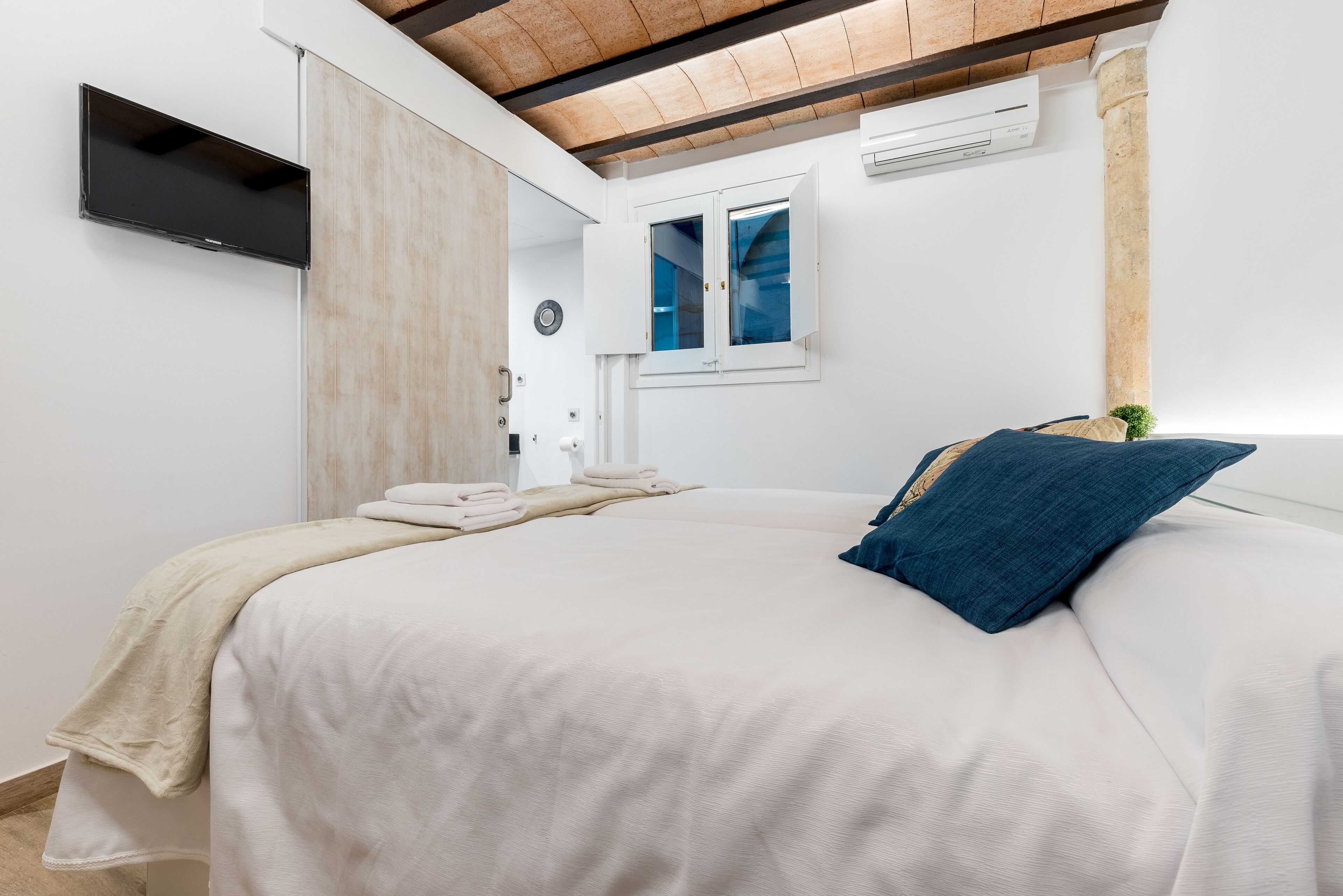 A bedroom with a bed, a flat-screen TV, a window, and a wooden ceiling beam.