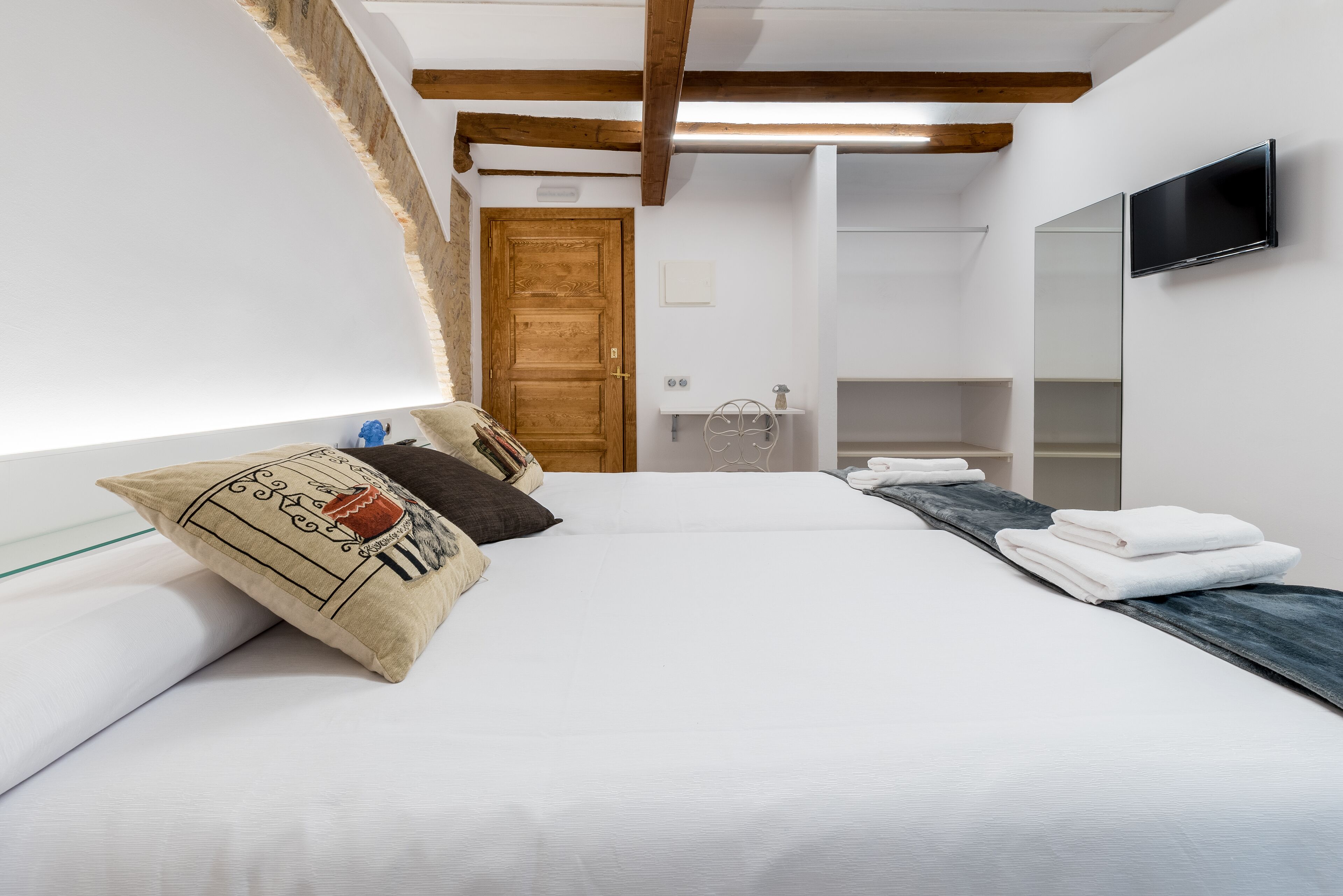 A modern bedroom with a large bed, wooden beams, a TV, and a bathroom visible in the background.