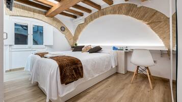 A bedroom with a bed, a desk, and a chair. There is a window with curtains and a wall-mounted air conditioner.