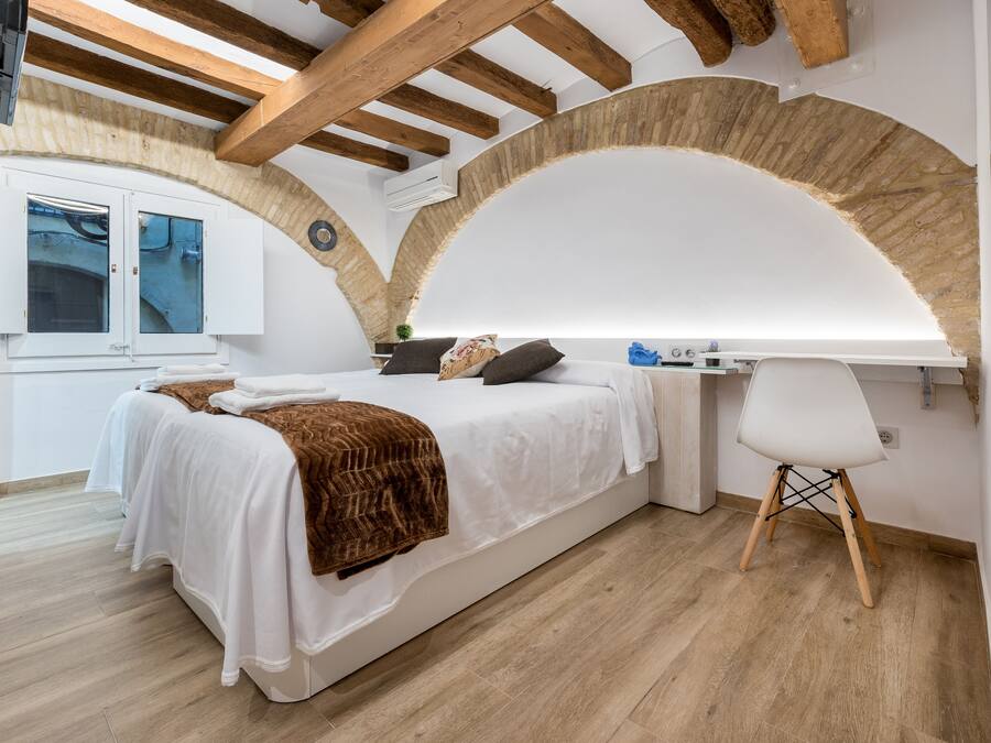 A bedroom with a bed, a desk, and a chair. There is a window with curtains and a wall-mounted air conditioner.