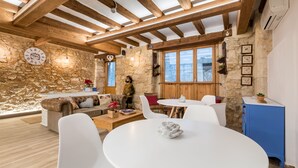 A rustic interior with stone walls, wooden beams, a round dining table, white chairs, and a cozy seating area with a sofa and cushions. - Hostal 977 (Tarragona)
