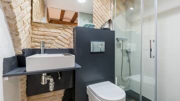 Een badkamer met een stenen muur, een witte wastafel, een zwart aanrecht, een toilet en een douchecabine van glas.