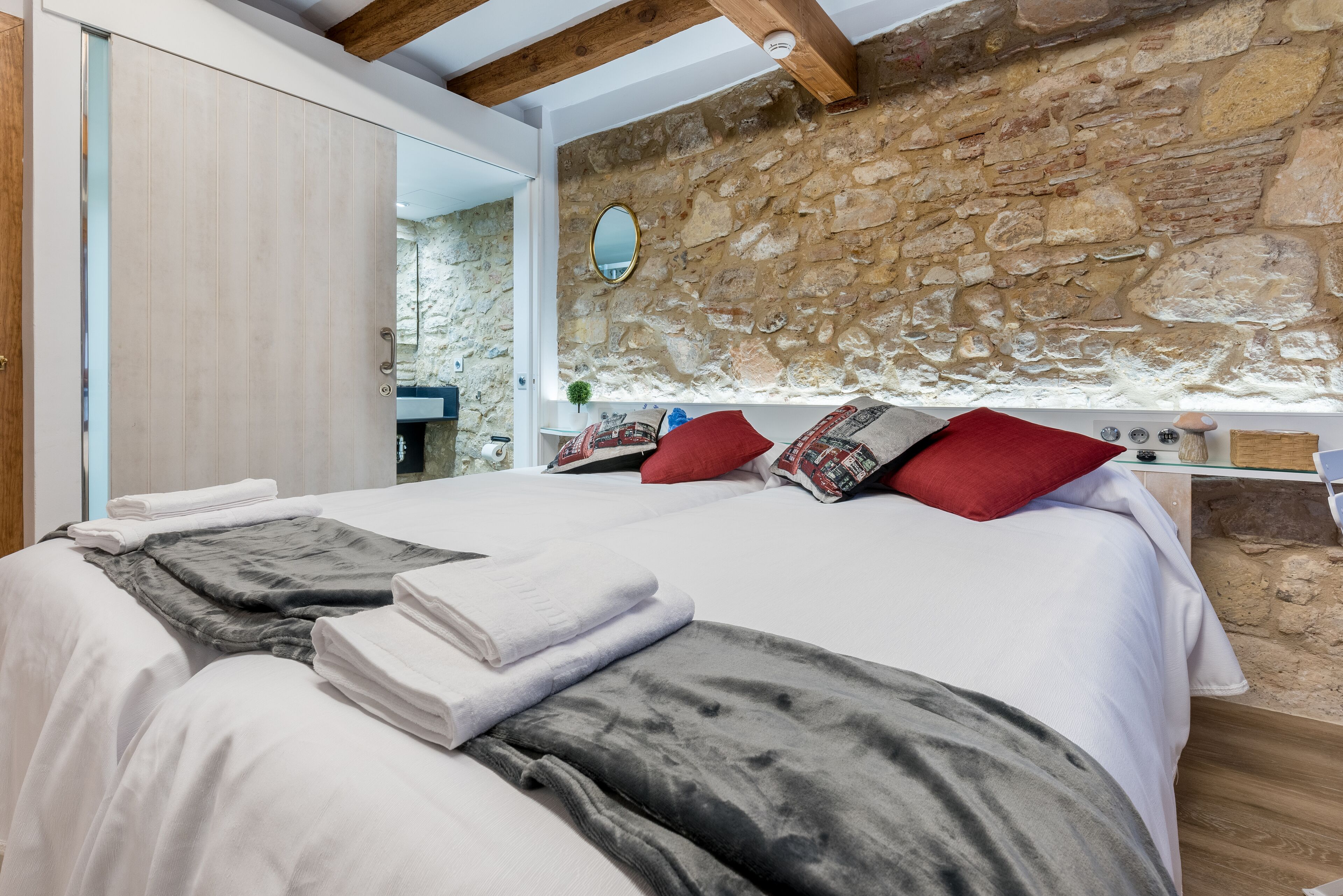 A bedroom with a stone wall, a bed with white linens, a red pillow, and a wooden ceiling.