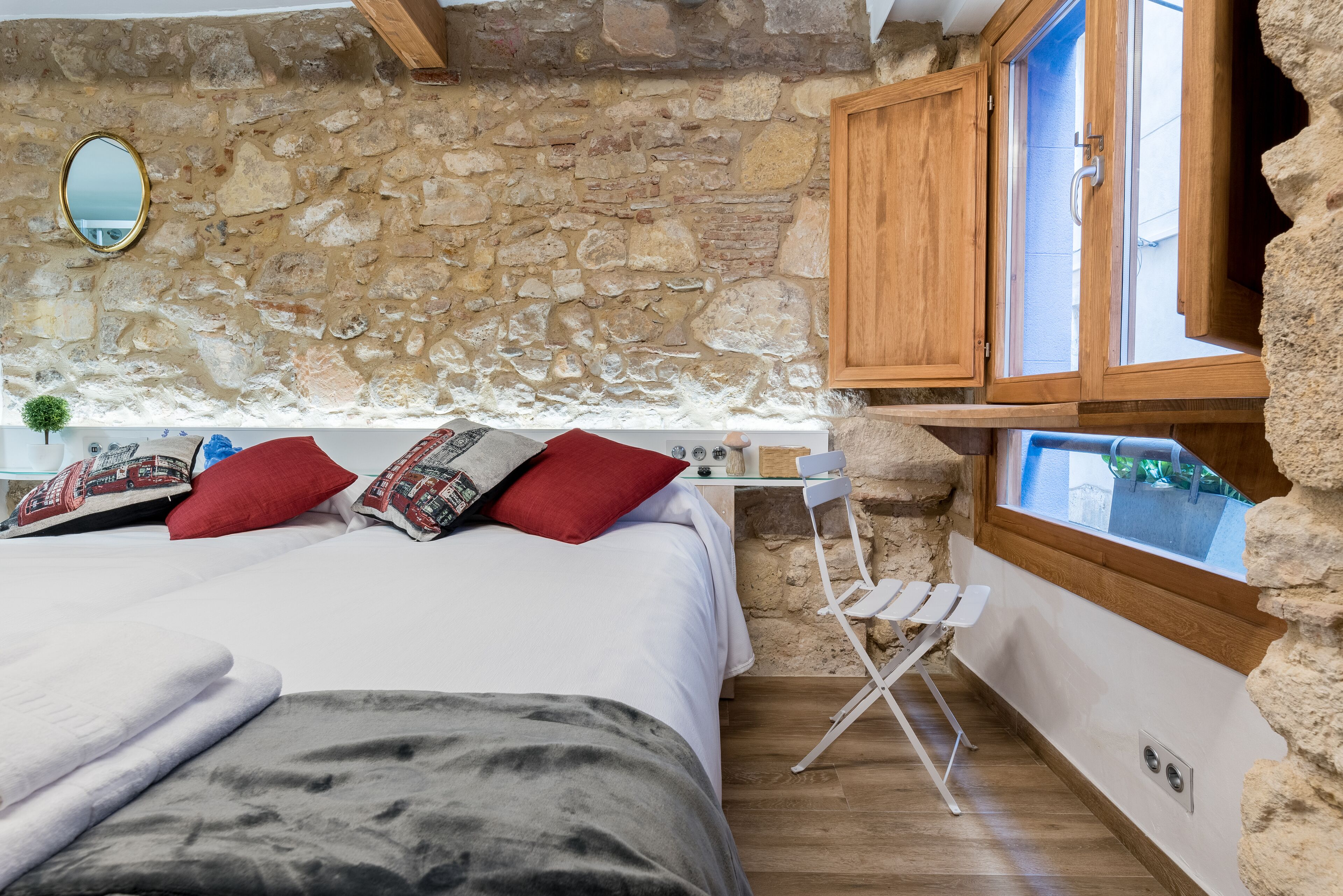 Un dormitorio con pared de piedra, armarios de madera, una cama con varias almohadas, una mesita y una ventana con vistas.