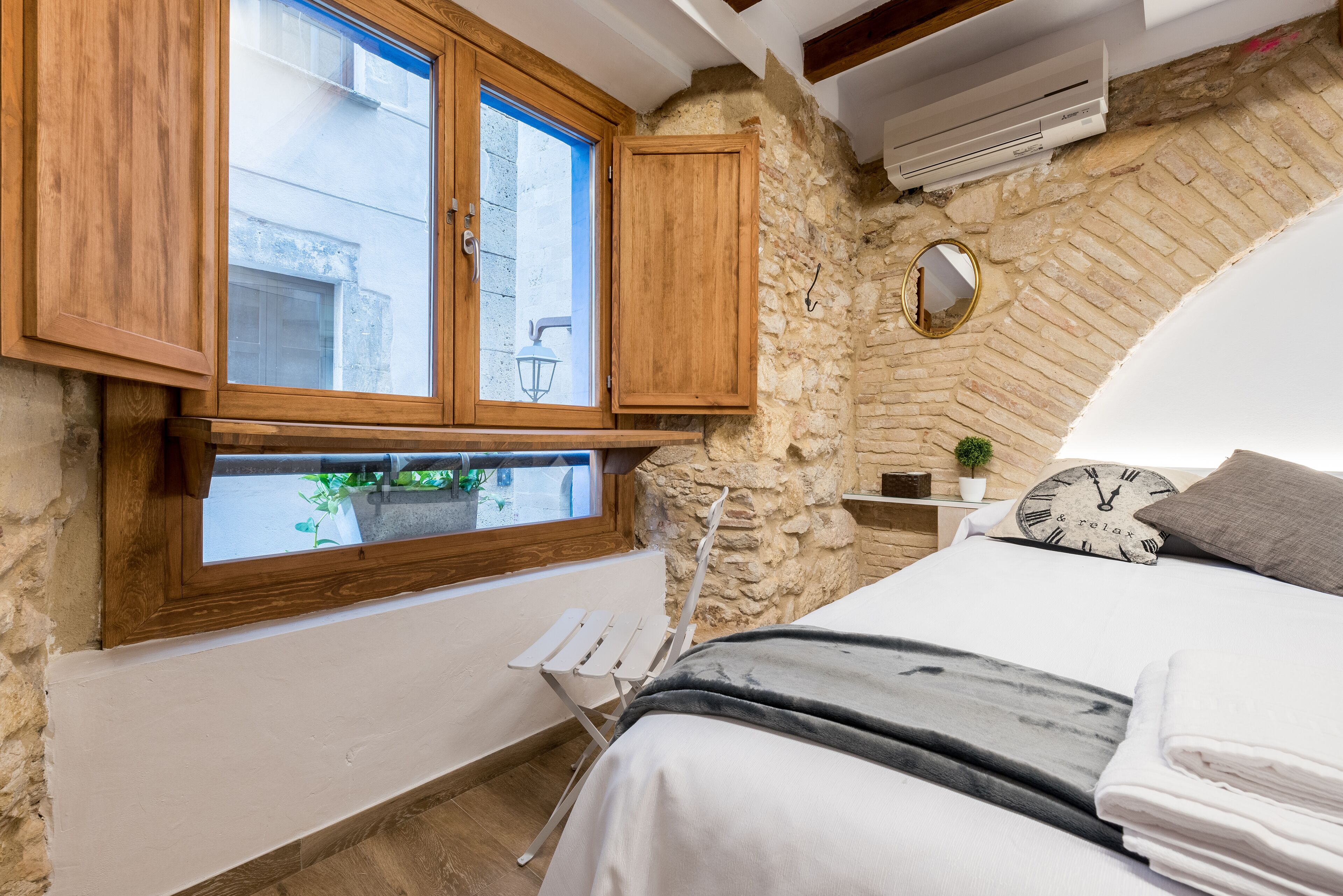 Un dormitorio con pared de piedra, armarios de madera, una cama con edredón blanco, un reloj y una pequeña planta en maceta.