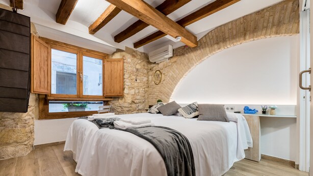 A bedroom with a stone wall, wooden beams, a bed with white and grey bedding, a window, and a small desk with a plant. - Hostal 977 (Tarragona)