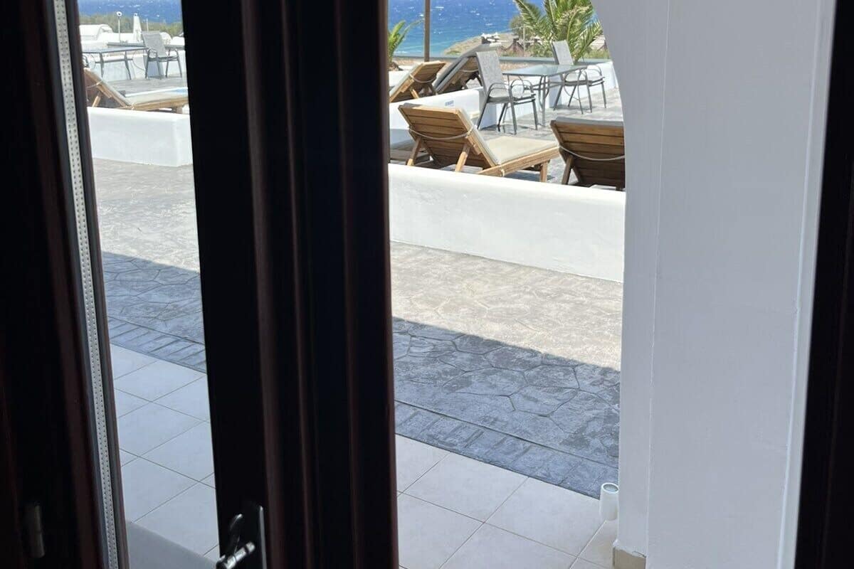 Vista a través de un ventanal a un patio con sillones de descanso y una sombrilla de paja, con vista a una playa y al océano.