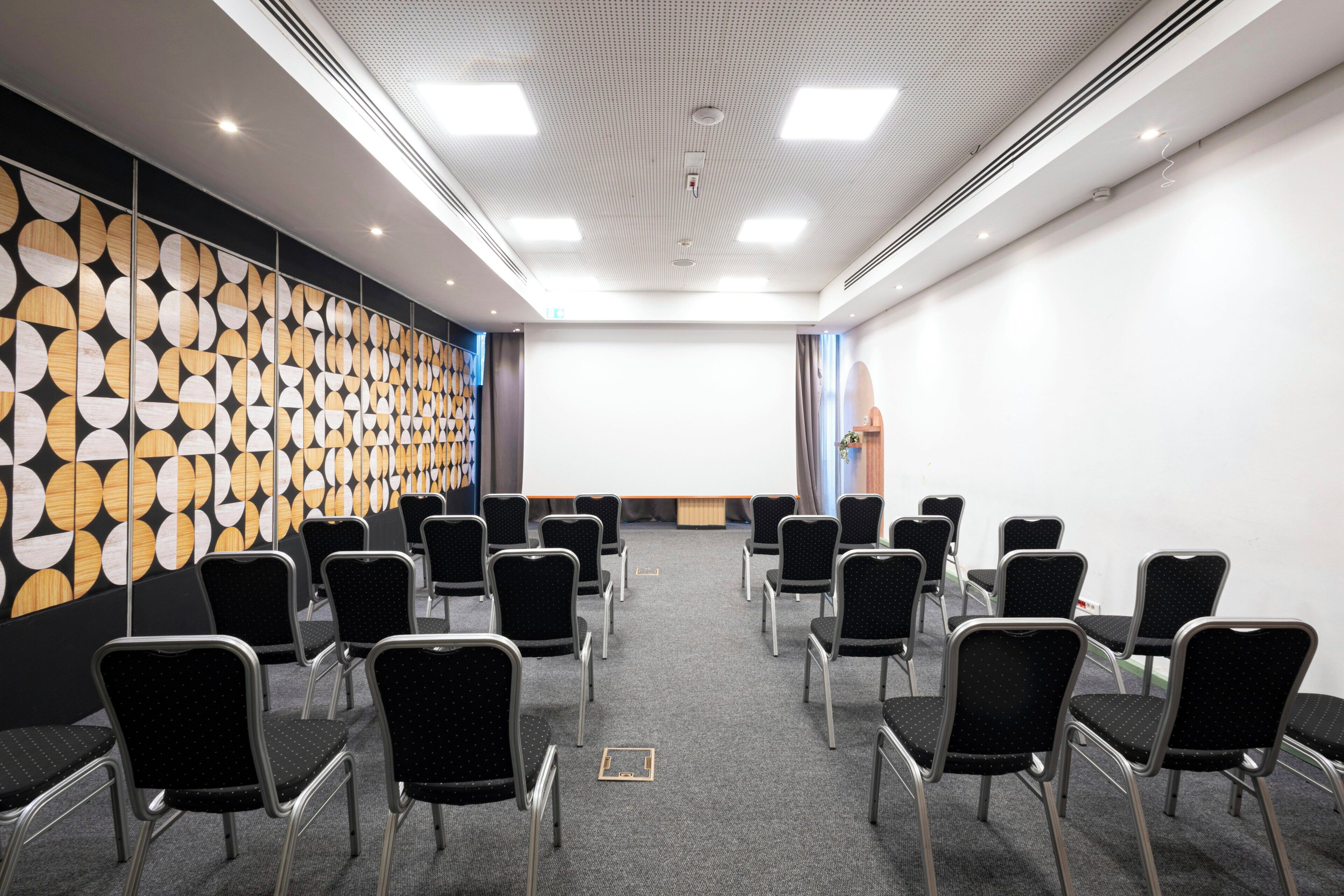 A conference room with a patterned wall, rows of chairs, and a projector screen.