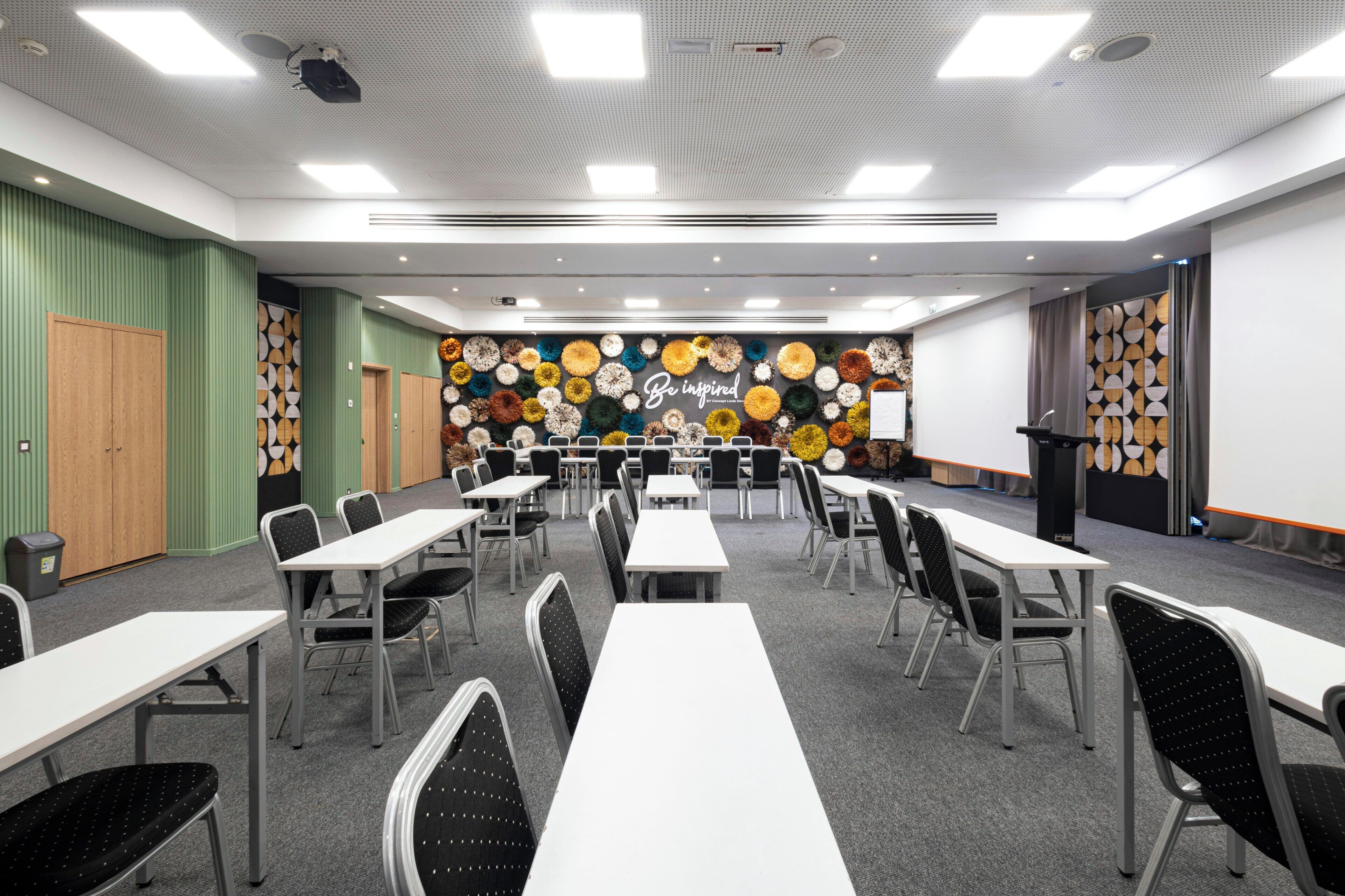 Uma sala de conferências com mesas, cadeiras, um projetor e um painel decorado com arranjos florais circulares.