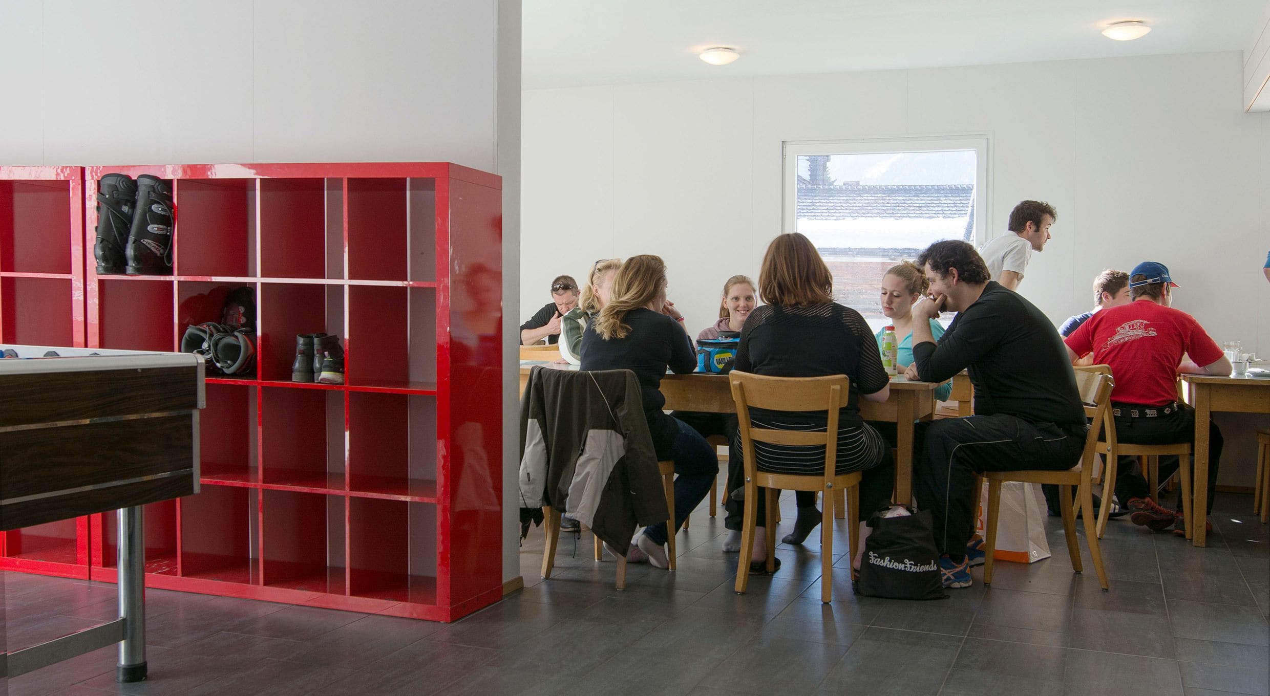 Un groupe de personnes est assis à des tables dans une pièce où l’on aperçoit une étagère rouge et un piano.