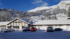 Un edificio cubierto de nieve con zona de estacionamiento para coches, rodeado de árboles y montañas nevadas.