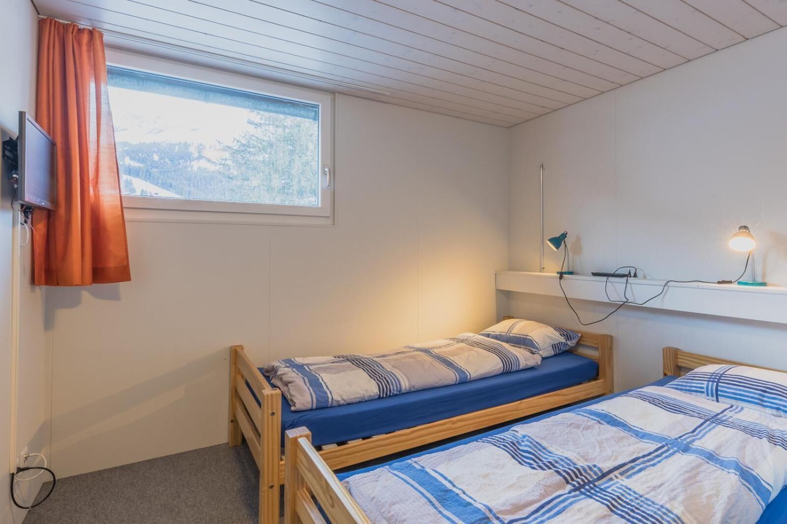A small, single-bed room with a window, a desk with a lamp, and a shelf with a lamp.