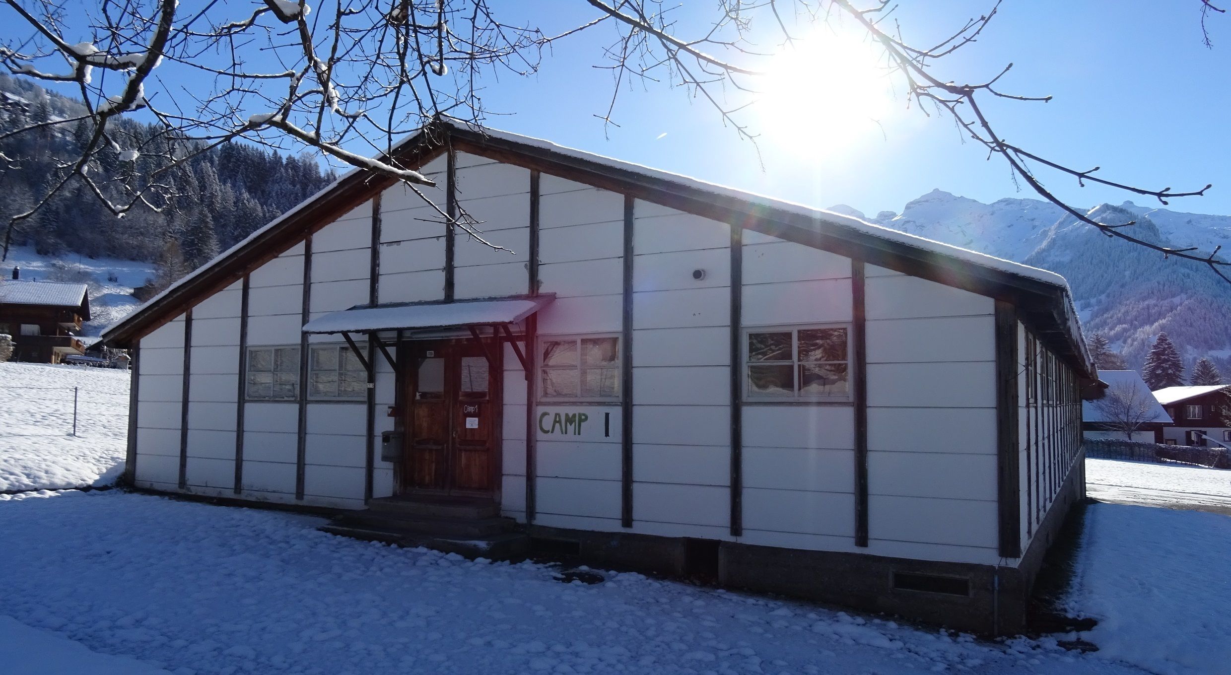 A white building labeled "Camp I" with a wooden door and windows, surrounded by snow and mountains.