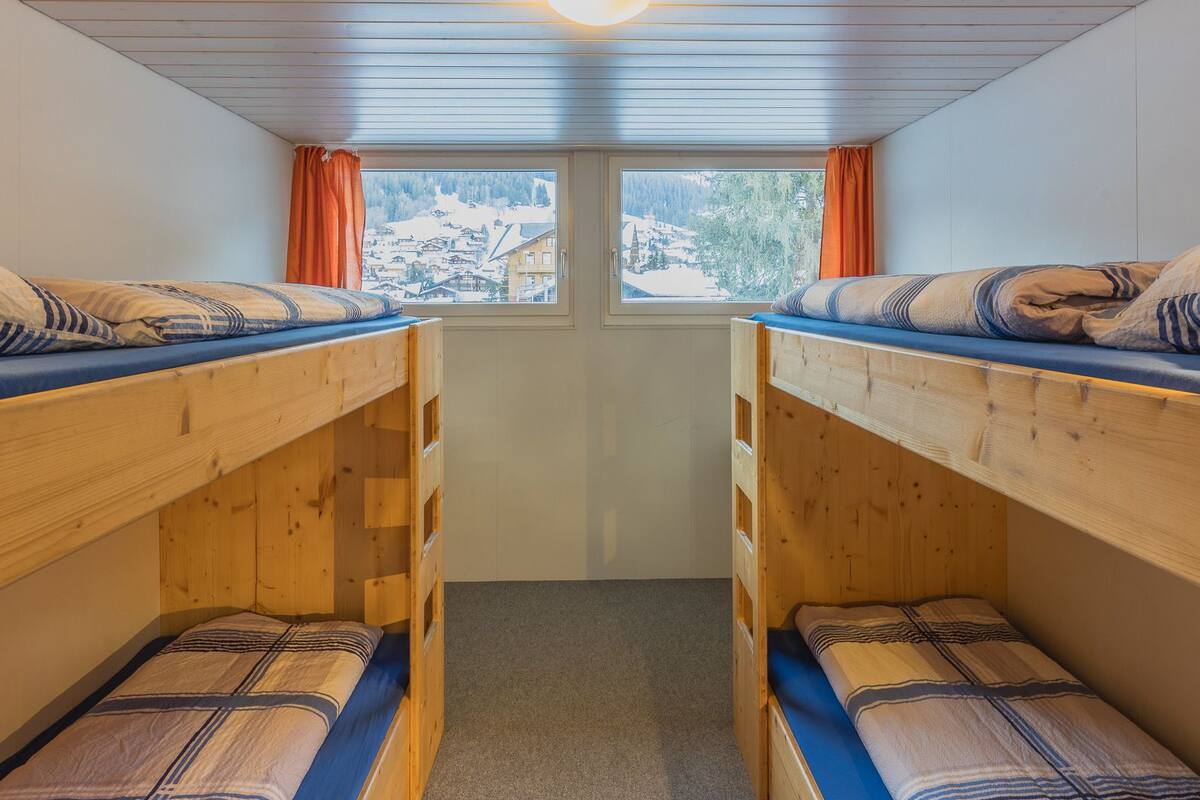 Quarto em dormitório com duas beliches, uma janela com cortinas e vista para uma paisagem nevada.